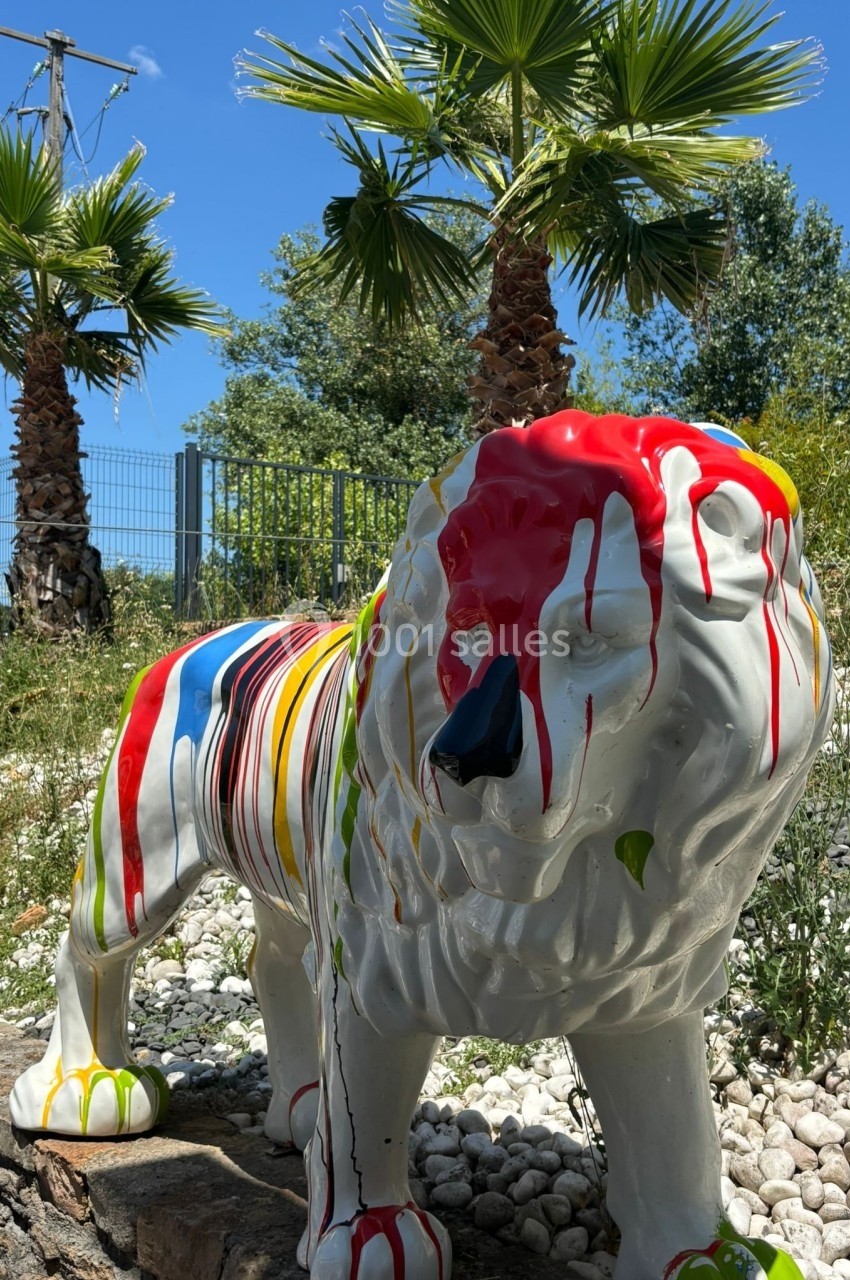 Statue de lion blanc recouverte de peinture multicolore, entourée de palmiers et de végétation sous un ciel bleu.