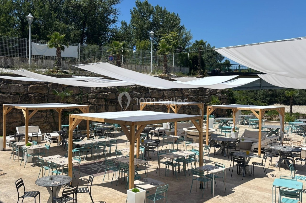 Terrasse extérieure avec tables et chaises sous des pergolas en bois et voiles d'ombrage, entourée de verdure.