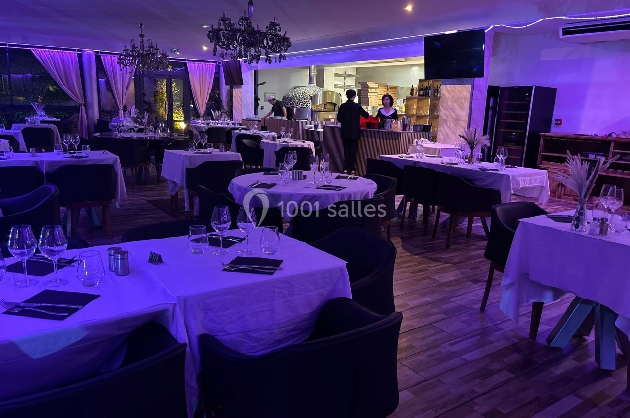 Salle de restaurant élégante avec tables dressées, éclairage tamisé violet et vue sur une cuisine ouverte.