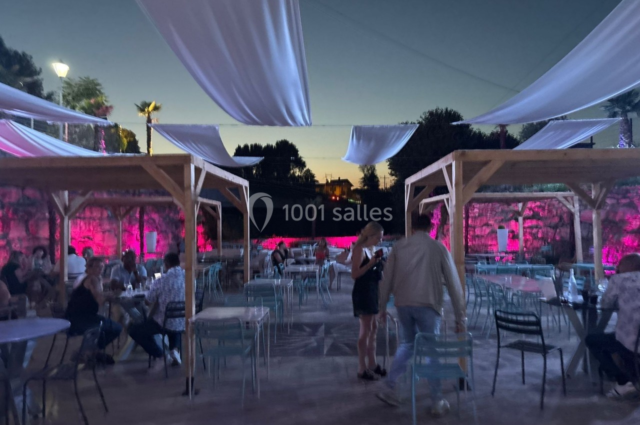 Terrasse extérieure en soirée avec des tables occupées, des voiles suspendues et un éclairage rose en arrière-plan.