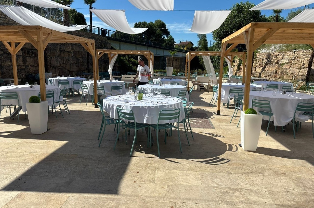 Terrasse extérieure aménagée avec des tables rondes dressées sous des pergolas en bois et des voiles blanches.