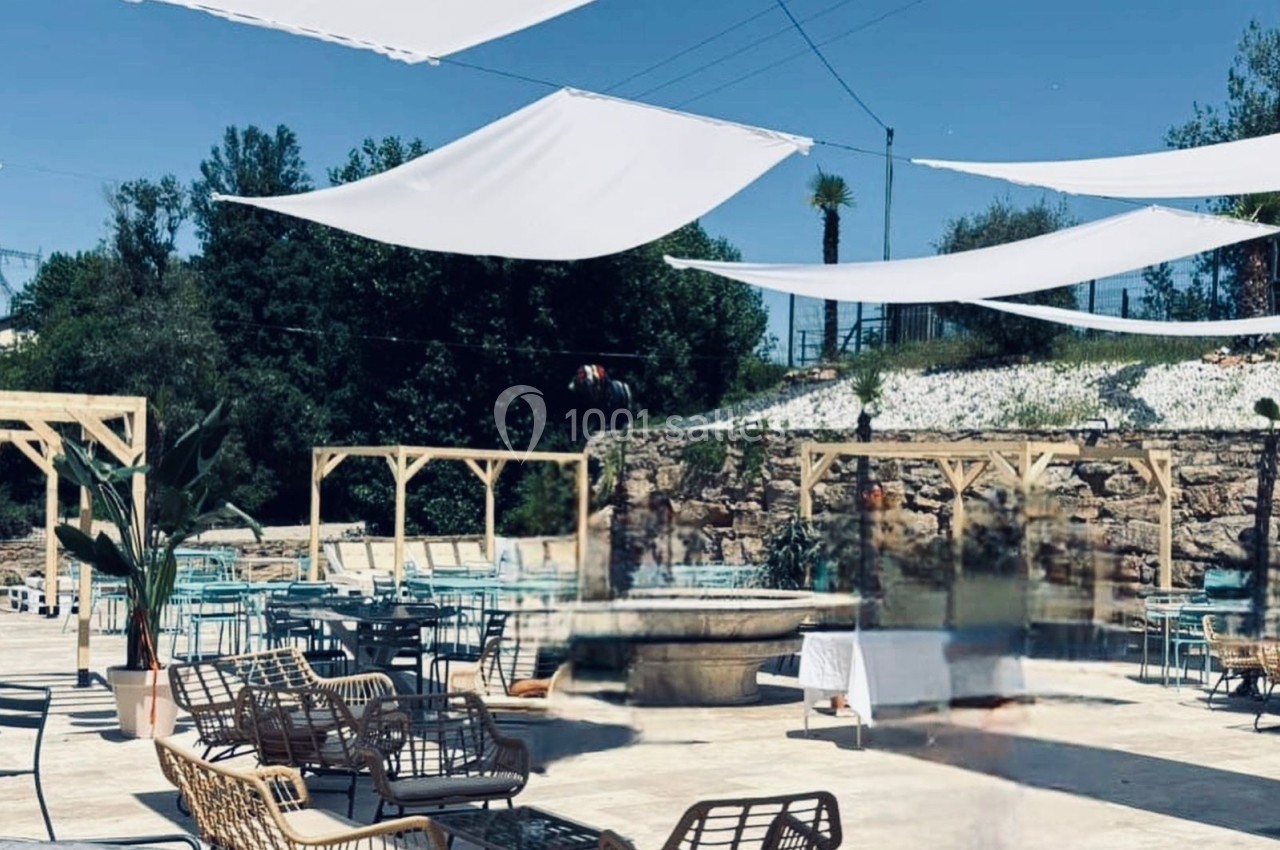 Terrasse extérieure avec des voiles d'ombrage blanches, mobilier en rotin et tables près d'une fontaine centrale.