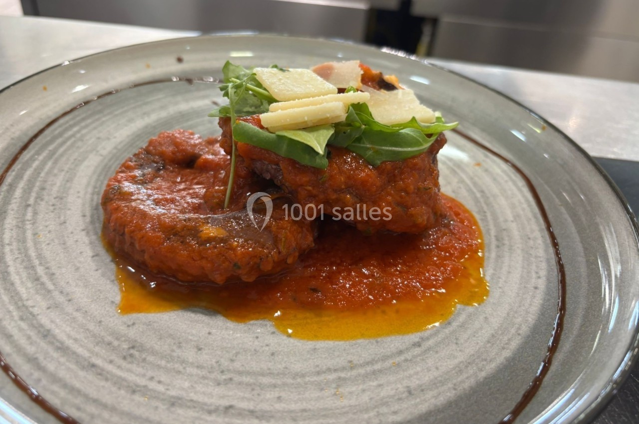 Assiette contenant des tranches de viande nappées de sauce tomate, garnies de roquette et de copeaux de fromage.