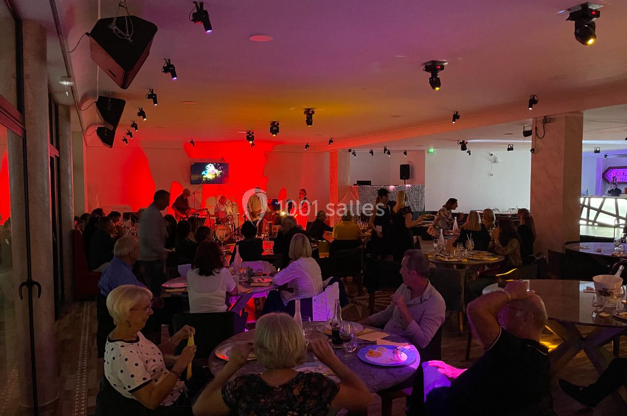 Salle de restaurant animée avec des convives attablés et un groupe de musiciens jouant sur une scène éclairée en rouge.