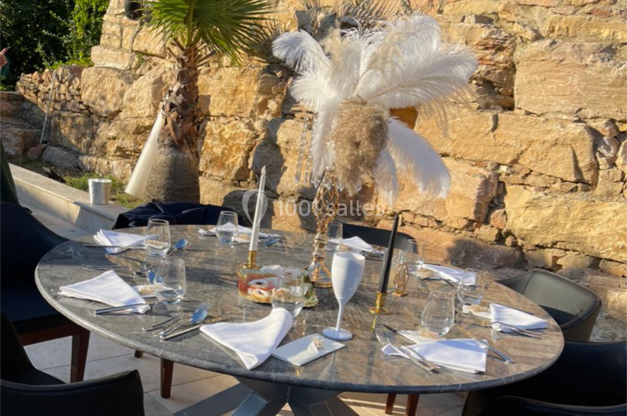 Table ronde dressée avec vaisselle élégante, bougies et décoration à plumes, en extérieur devant un mur de pierre.