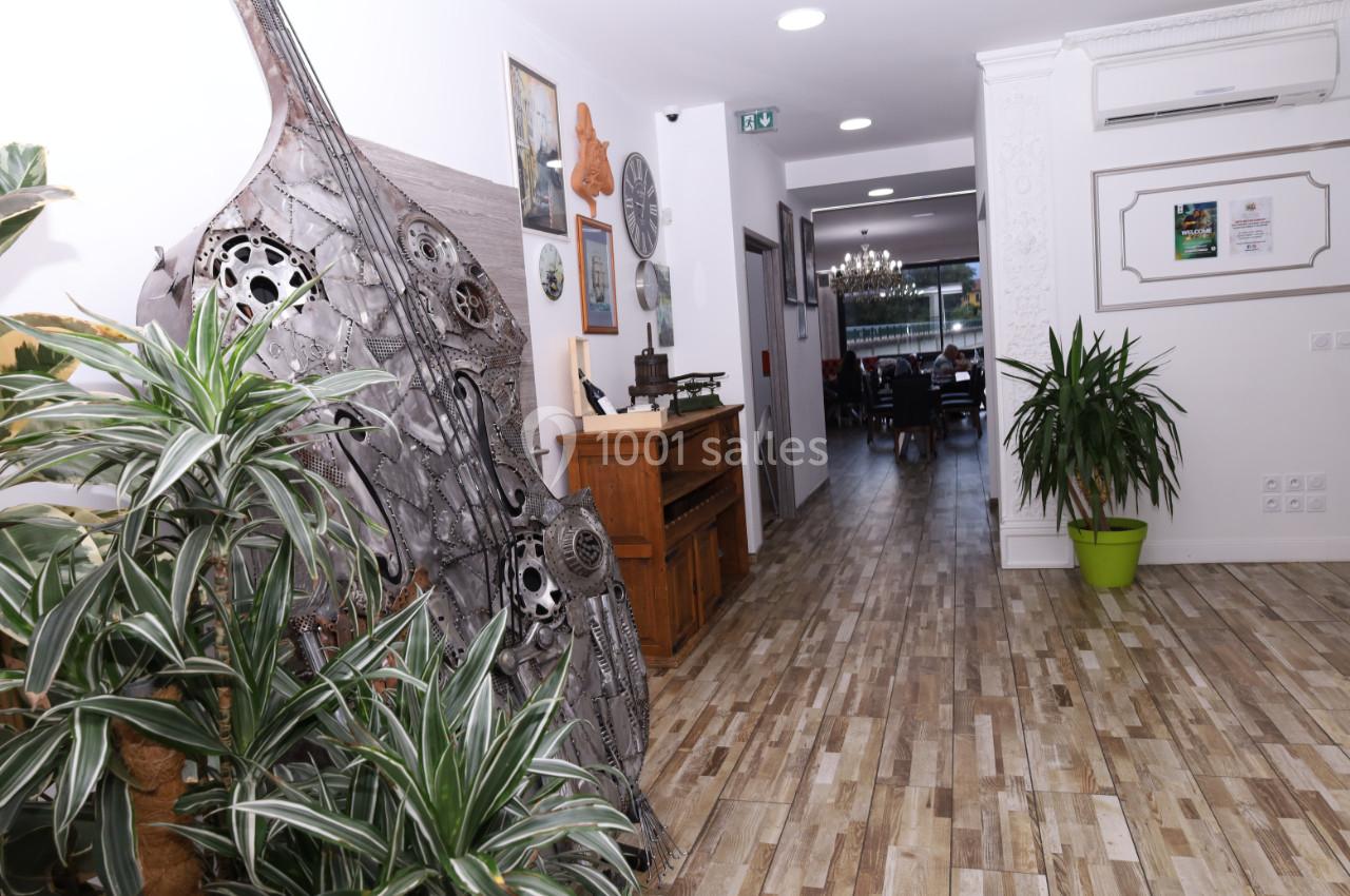 Couloir lumineux avec sol en bois, plantes vertes, sculptures métalliques et vue sur une salle à manger au fond.