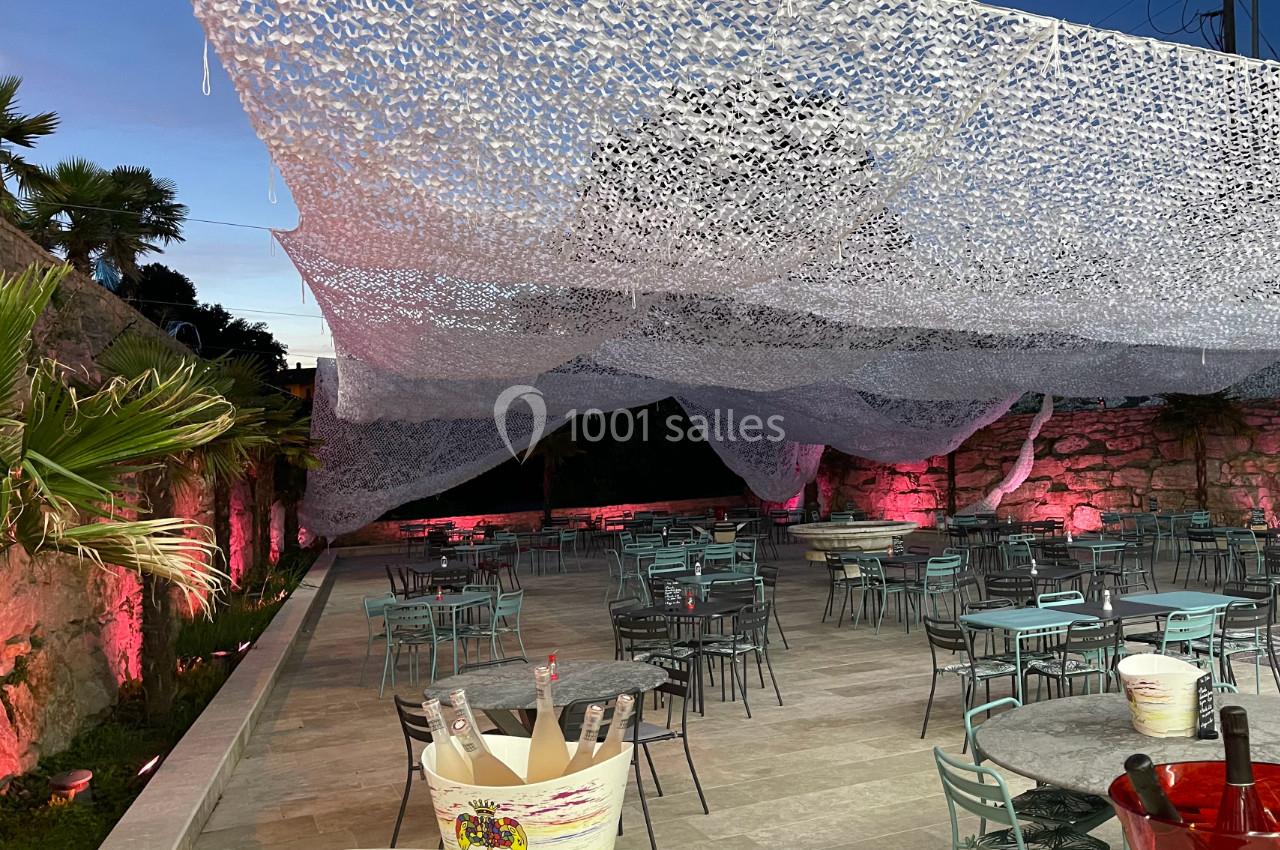 Terrasse extérieure aménagée avec des tables et chaises, décorée de filets suspendus et éclairée par des lumières roses.