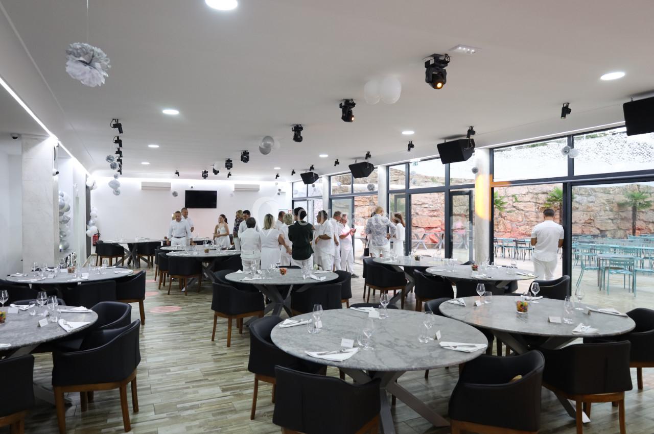 Salle de réception lumineuse avec tables dressées, invités en tenue blanche et grandes baies vitrées donnant sur une…