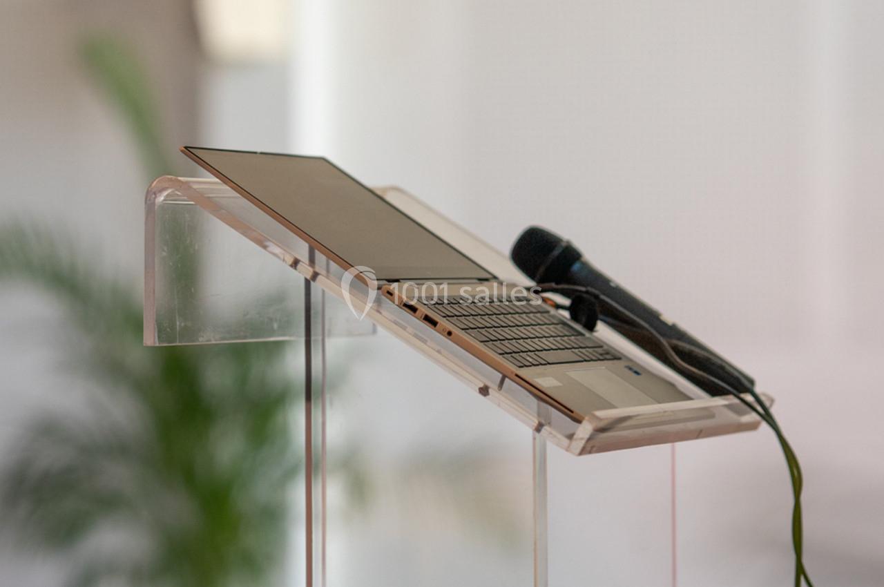 Ordinateur portable ouvert et microphone posé sur un pupitre transparent, avec une plante floue en arrière-plan.
