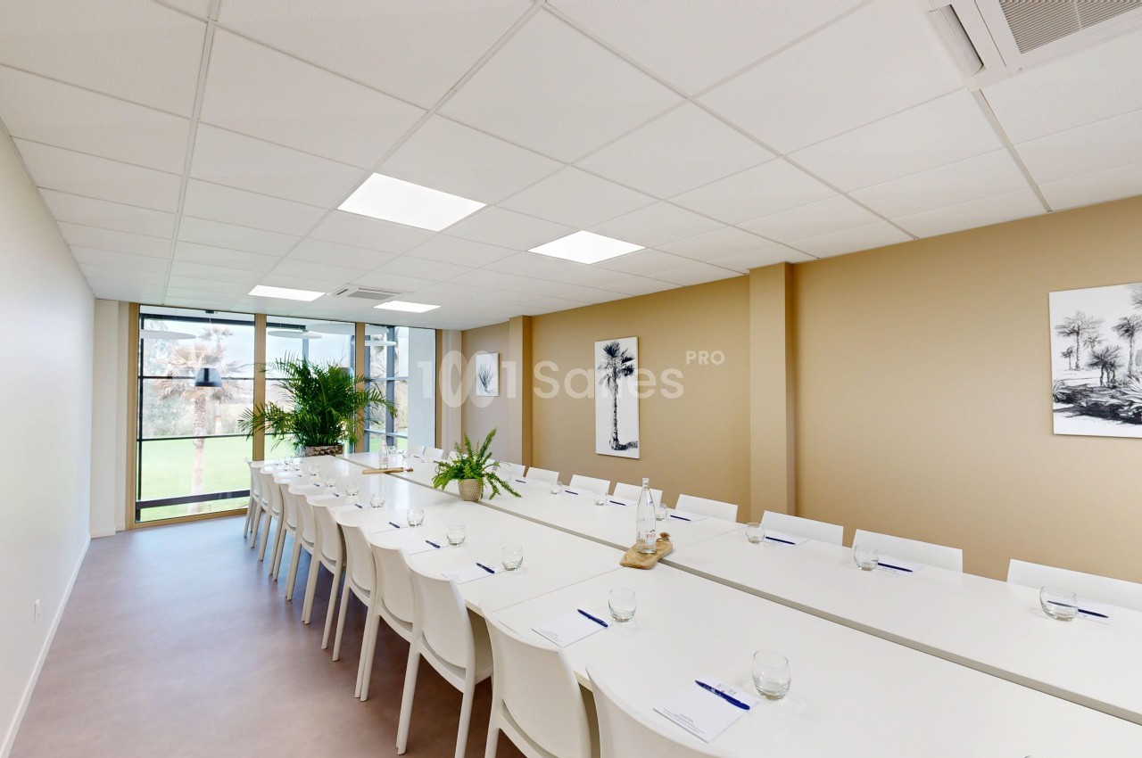 Salle de réunion lumineuse avec de longues tables blanches alignées, chaises, plantes et vue sur l'extérieur.