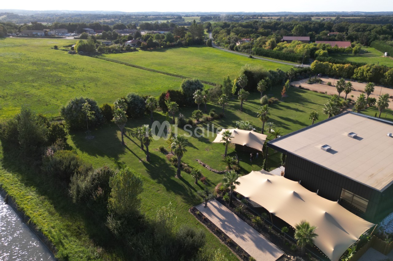 Vue aérienne d'un espace vert avec un bâtiment moderne, des arbres, des toiles d'ombrage et des champs en arrière-plan.
