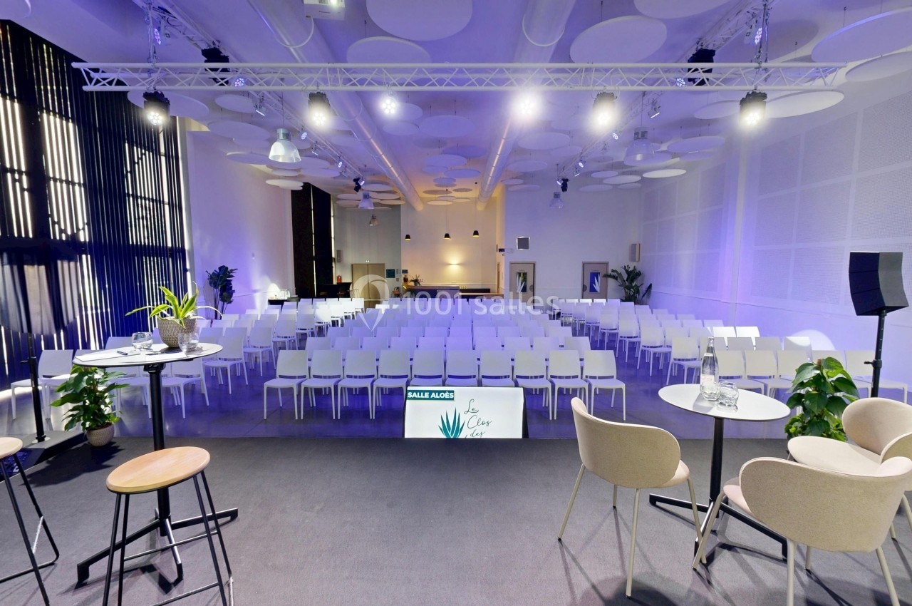 Salle de conférence moderne avec chaises blanches alignées, scène équipée de tabourets et tables, éclairage tamisé.