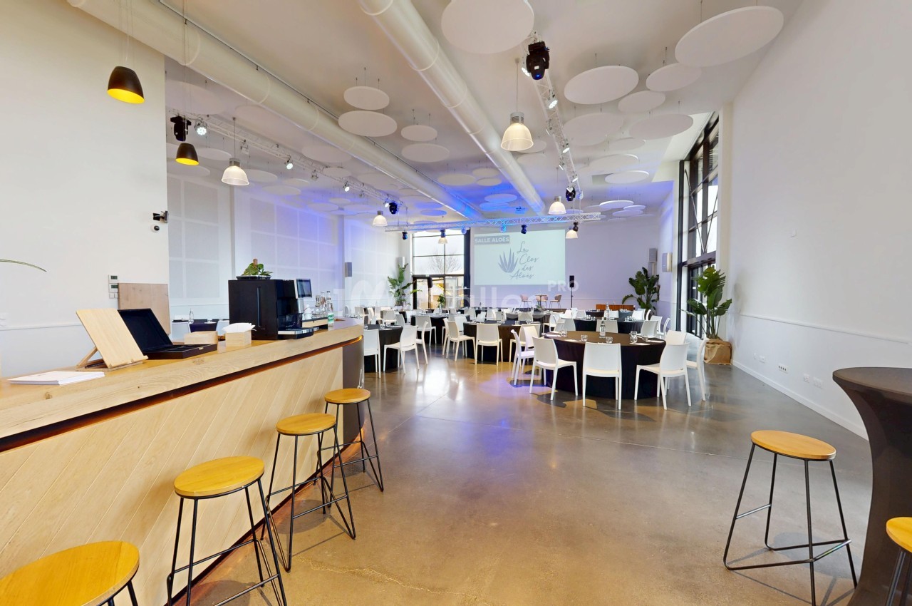 Salle lumineuse aménagée pour un événement, avec tables rondes, chaises blanches et un comptoir en bois au premier plan.