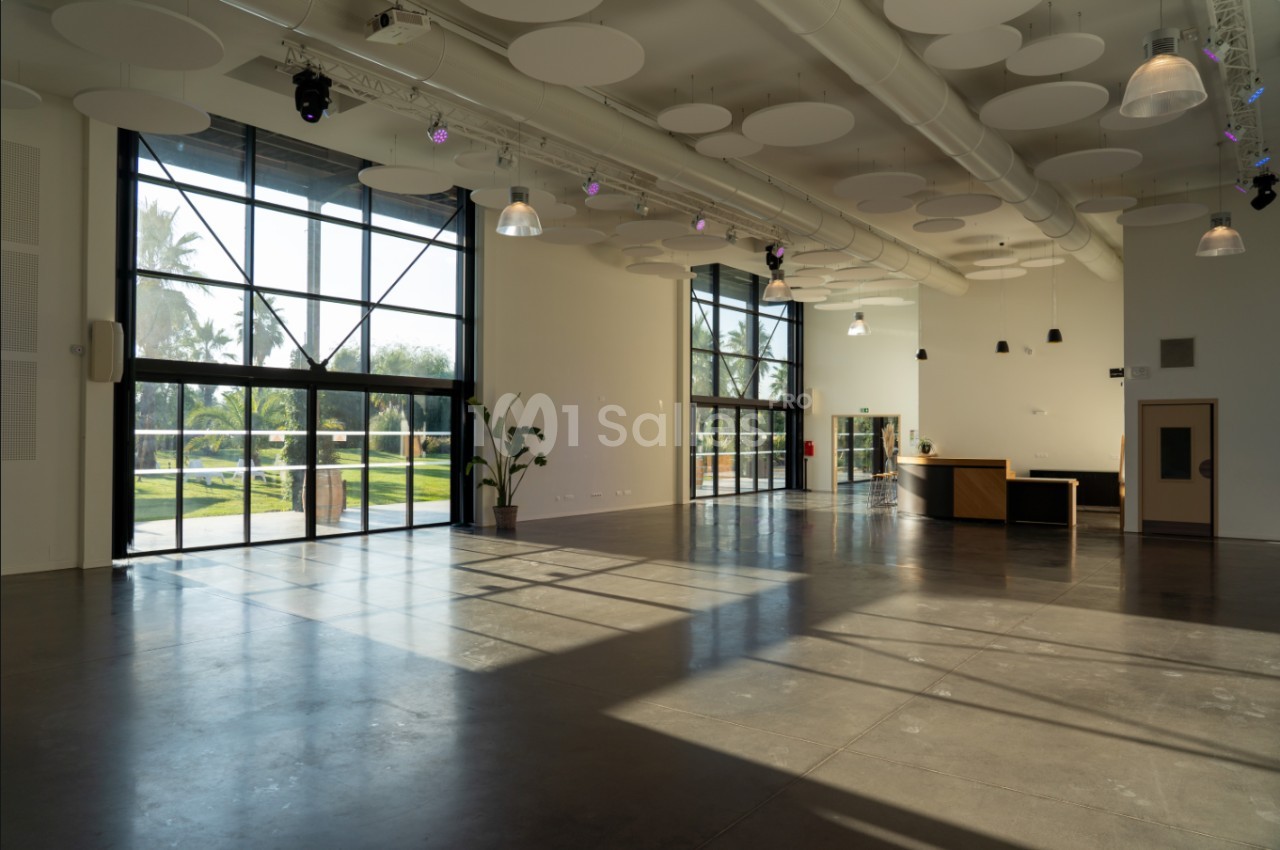 Salle lumineuse avec de grandes baies vitrées, un sol en béton poli, des plantes et un comptoir d'accueil.
