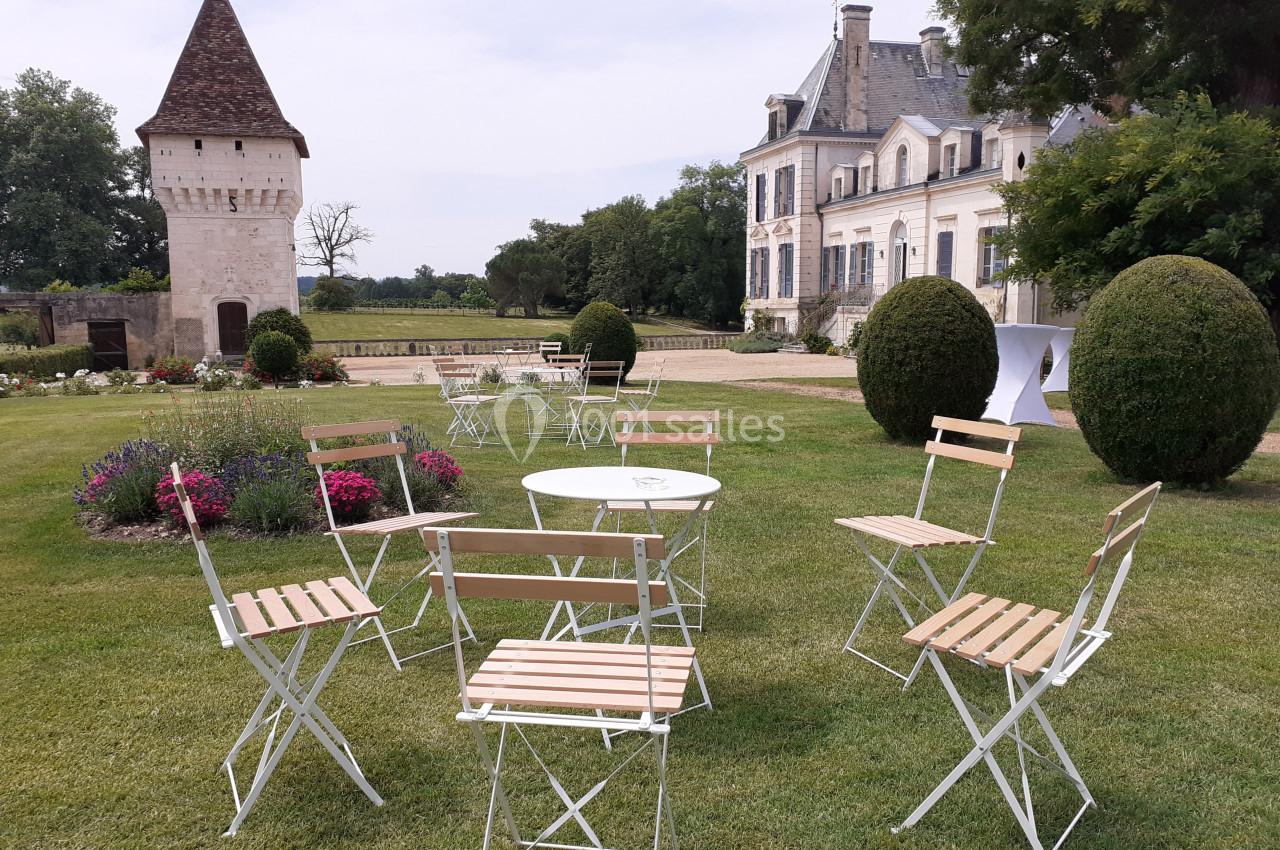 Chaises et tables blanches disposées sur une pelouse devant un manoir et une tour, entourés de fleurs et d'arbustes.