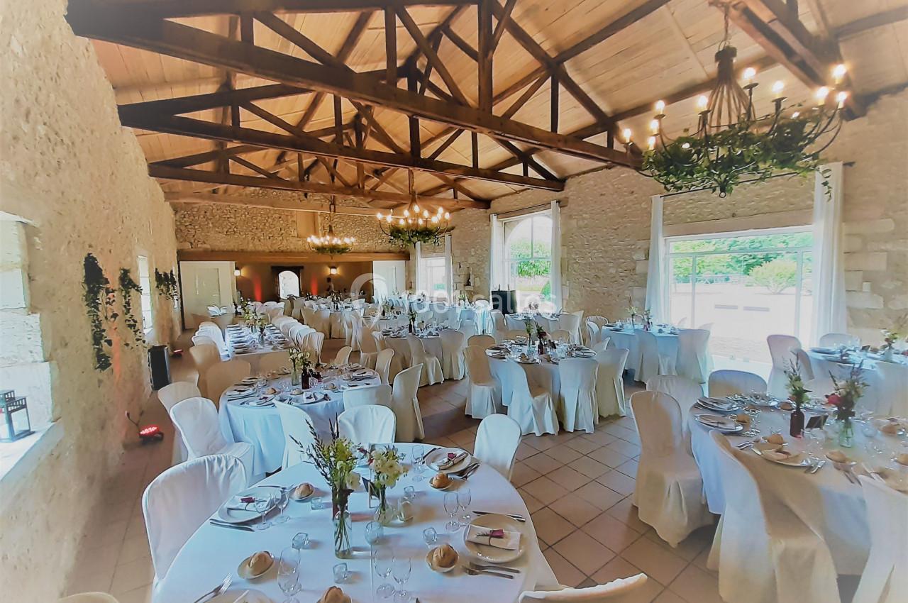 Salle de réception lumineuse avec tables rondes dressées, chaises blanches et poutres apparentes en bois.