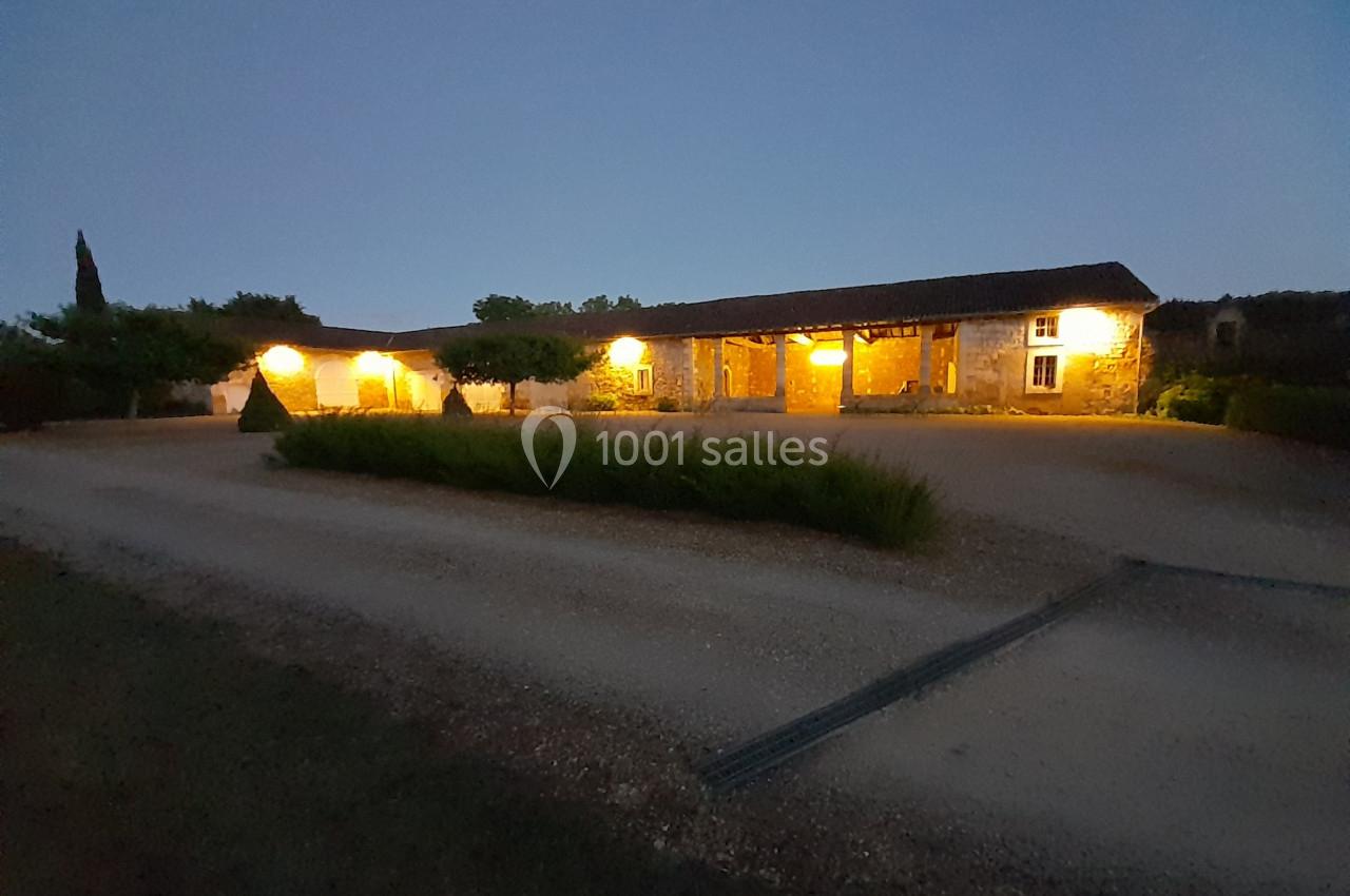 Bâtiment en pierre éclairé par des lumières extérieures, entouré de végétation, au crépuscule.