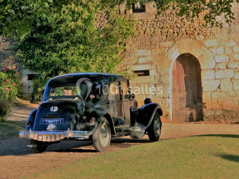 Voiture ancienne noire stationnée devant une bâtisse en pierre, entourée de verdure et baignée de lumière.