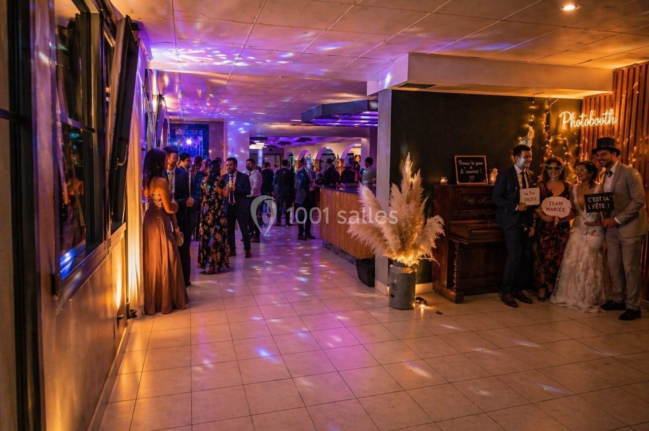 Salle de réception illuminée avec des invités debout et un coin photobooth décoré de pampas et d'accessoires festifs.