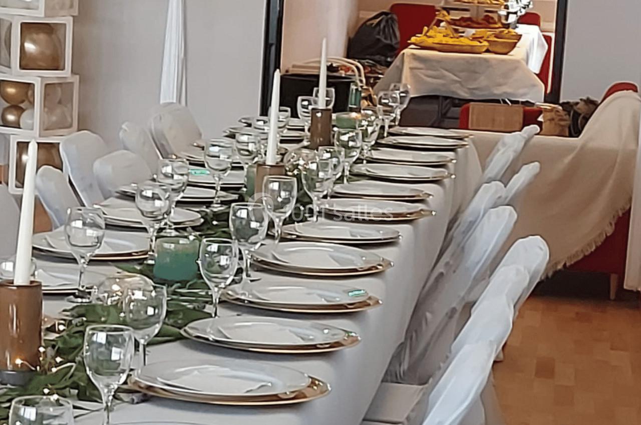 Table dressée avec nappes blanches, assiettes, verres et bougies, dans une salle avec buffet visible en arrière-plan.