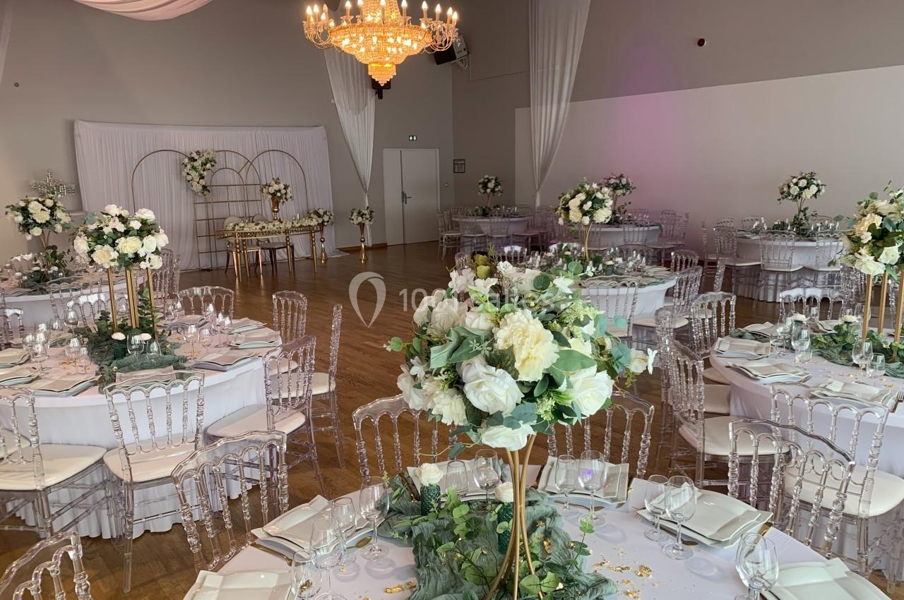 Salle de réception décorée avec des tables rondes, des chaises transparentes et des compositions florales blanches et vertes.