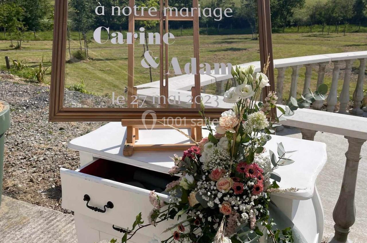 Panneau de bienvenue pour un mariage, posé sur un chevalet devant un paysage champêtre, avec un bouquet floral sur une table…