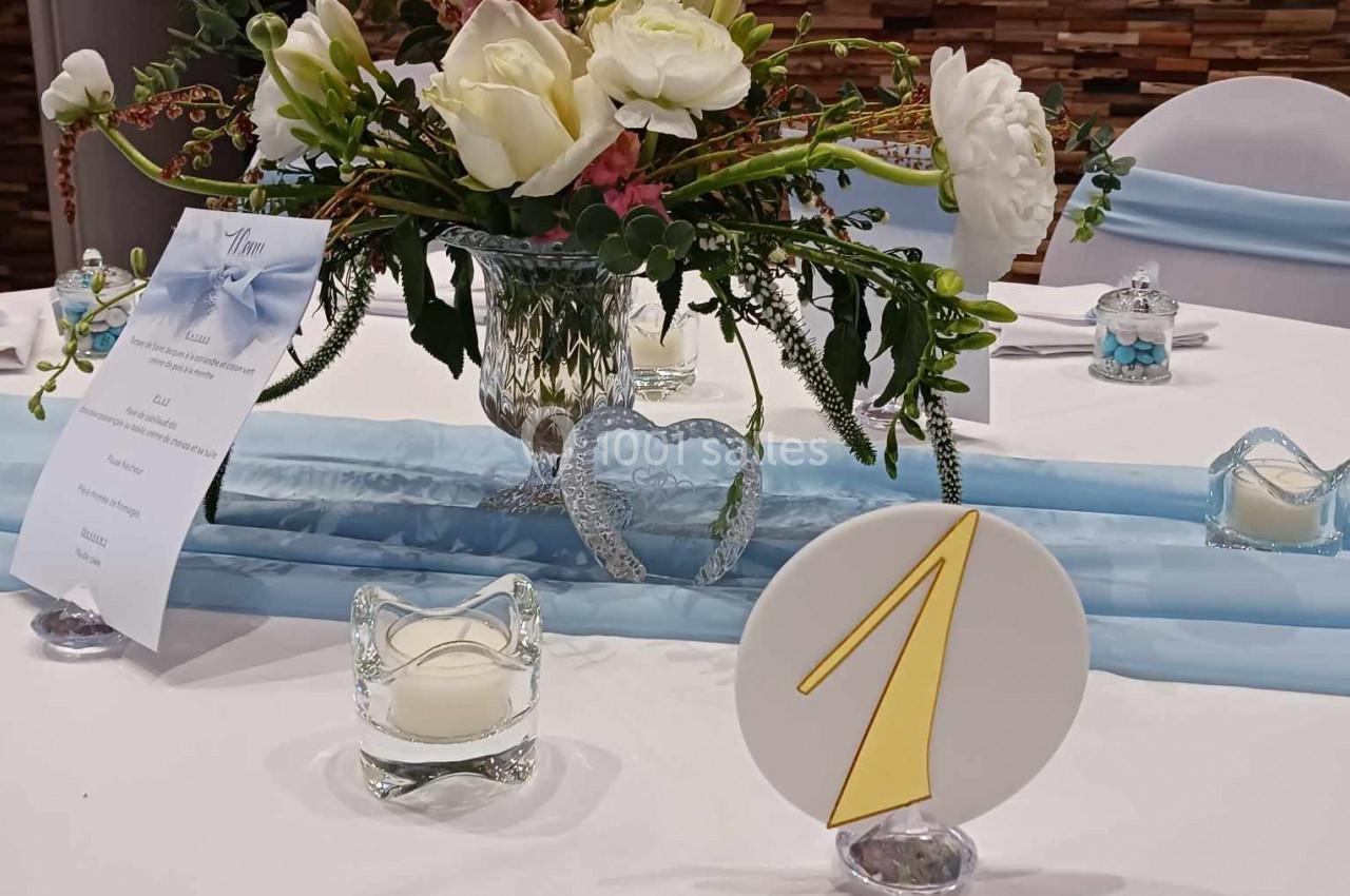 Centre de table avec bouquet de fleurs blanches et roses, numéro de table ’1’ et bougies sur une nappe blanche.