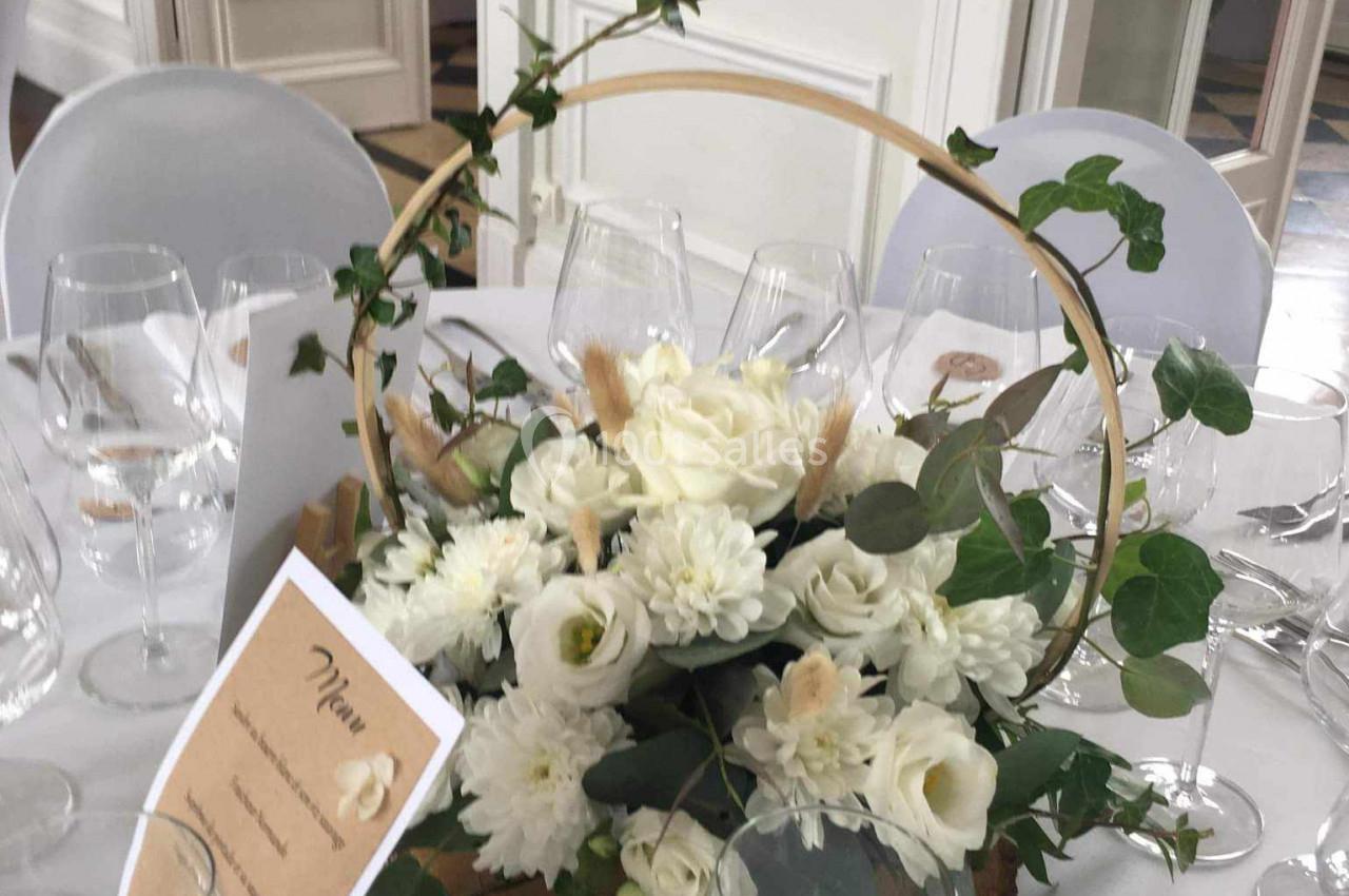 Centre de table floral avec roses blanches, feuillage et structure circulaire en bois, entouré de verres et d'un menu.