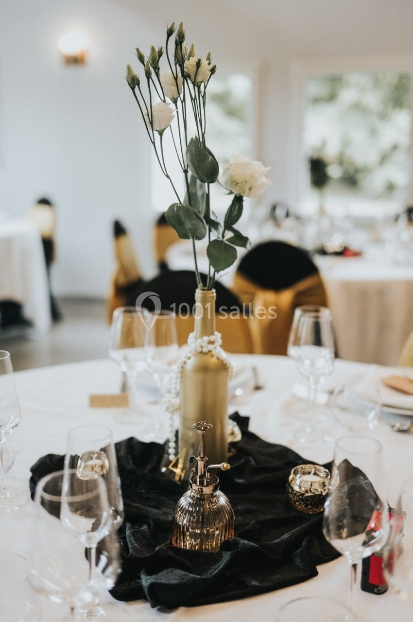 Centre de table élégant avec fleurs blanches dans une bouteille dorée, perles et décorations sur une nappe noire.