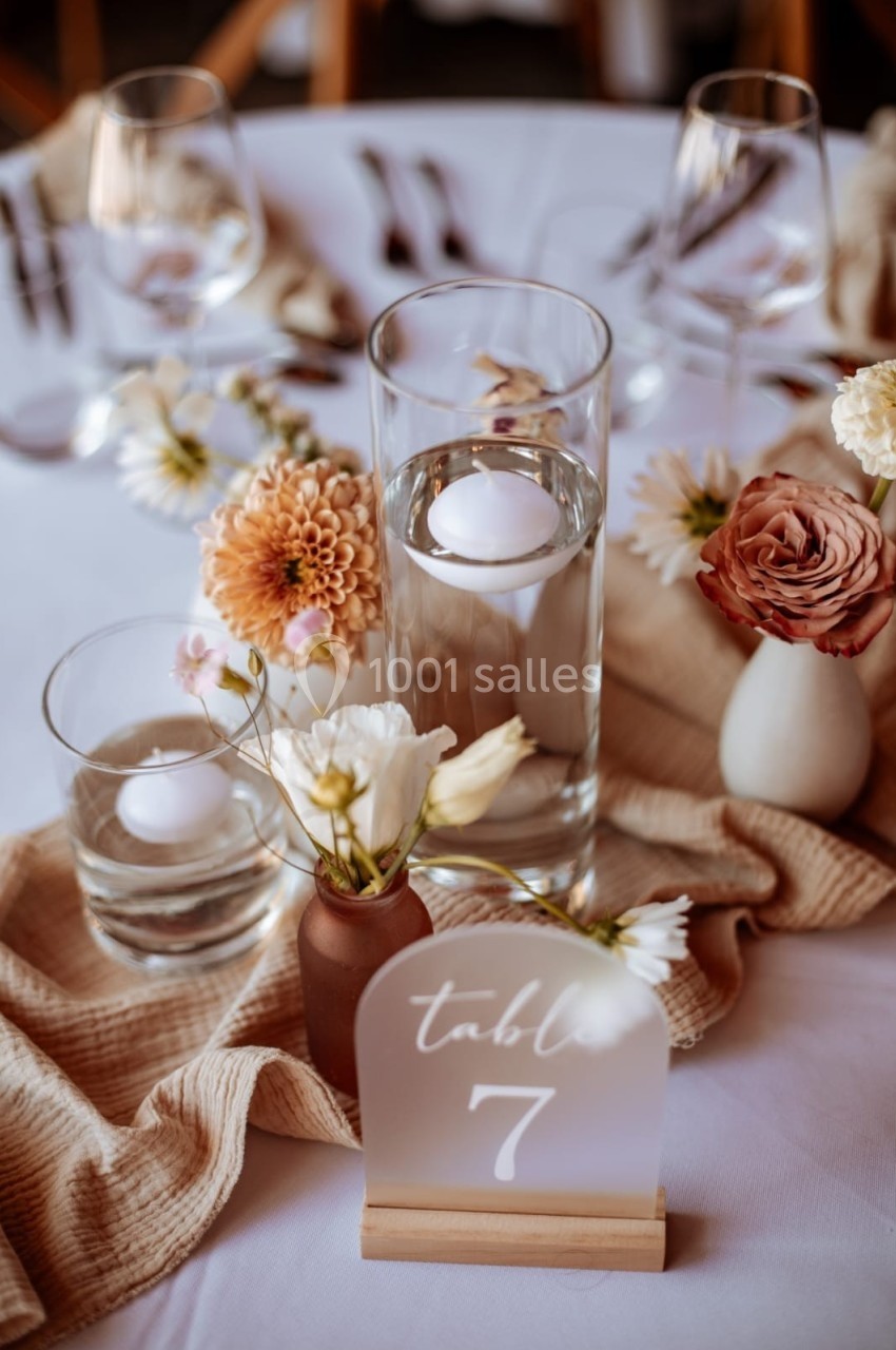 Centre de table avec fleurs pastel, bougies flottantes dans des vases en verre et panneau indiquant ’Table 7’.