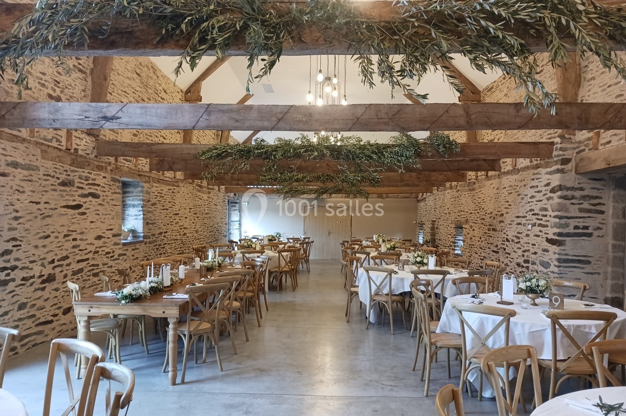 Salle de réception en pierre avec poutres apparentes, tables rondes et rectangulaires décorées pour un événement.