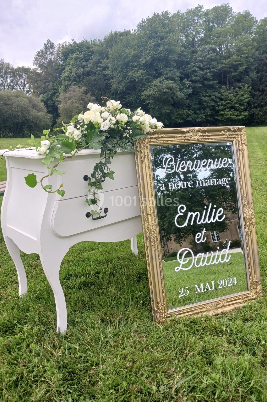 Commode blanche avec fleurs et miroir doré affichant un message de bienvenue pour un mariage en extérieur.