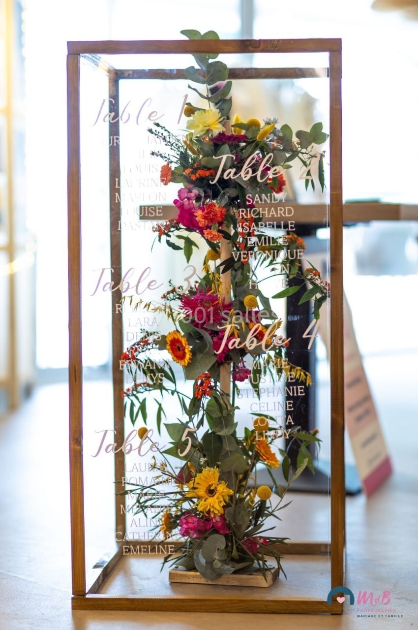 Plan de table de mariage dans une structure en bois et verre, décorée de fleurs colorées et de feuillage.