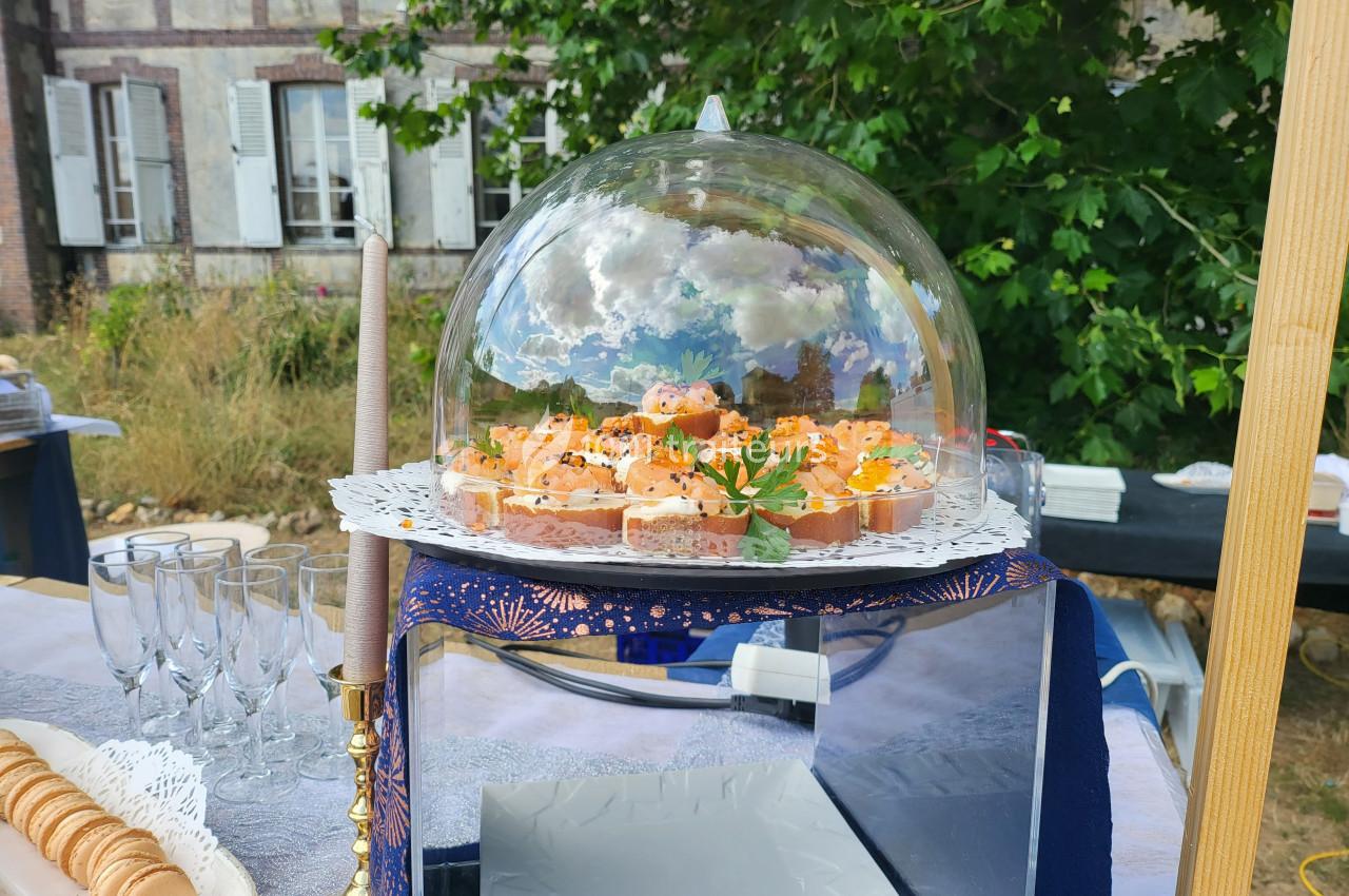 Plateau de bouchées apéritives sous une cloche en verre, présenté sur une table en extérieur près d'un bâtiment ancien.