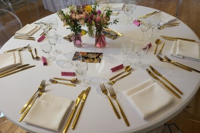 Table élégamment dressée avec nappes blanches, chaises assorties, chandeliers, fleurs roses et couverts dorés.