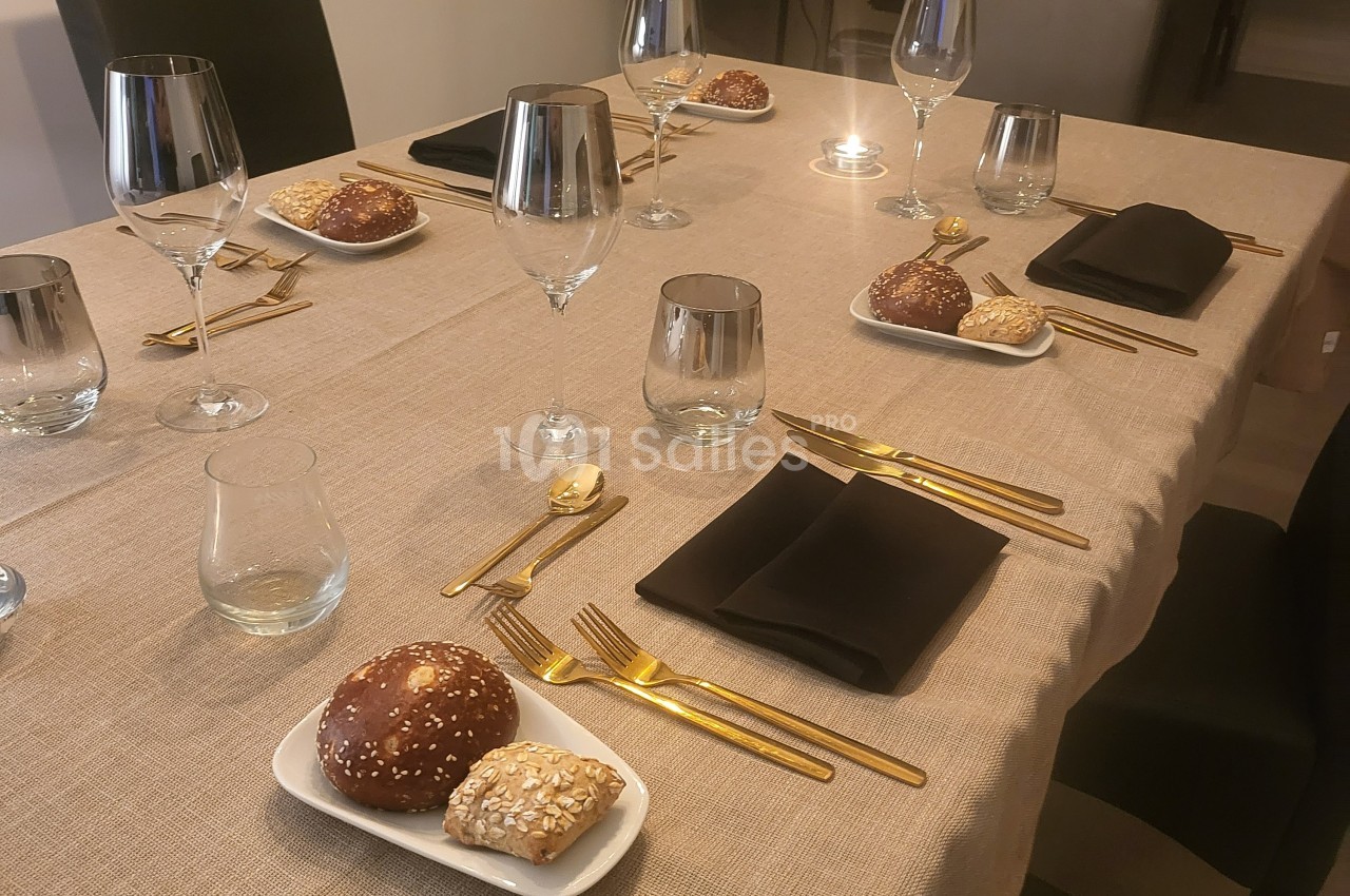 Table dressée avec une nappe beige, couverts dorés, serviettes noires, verres et assiettes contenant des petits pains.