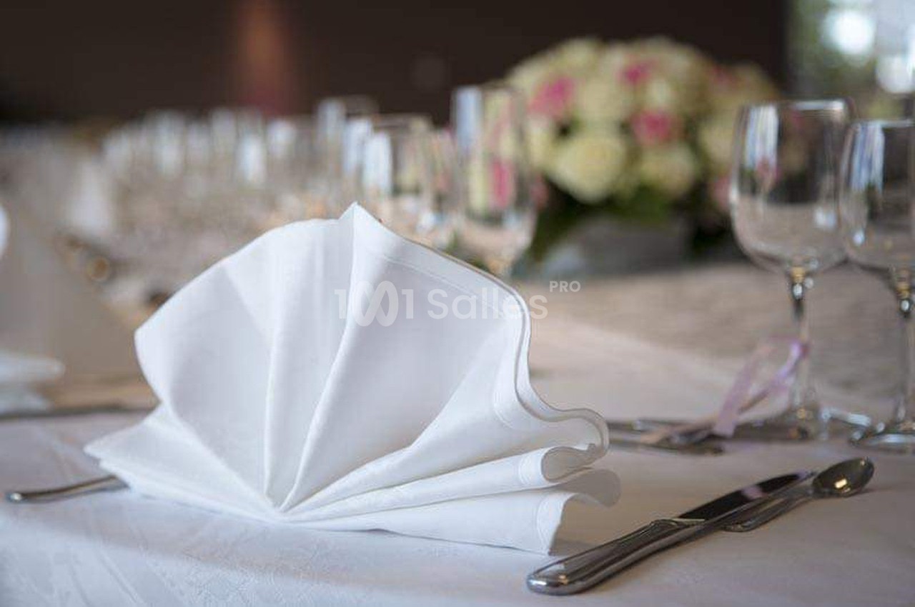 Serviette pliée en éventail sur une table dressée avec des verres, couverts et un centre de table floral en arrière-plan.