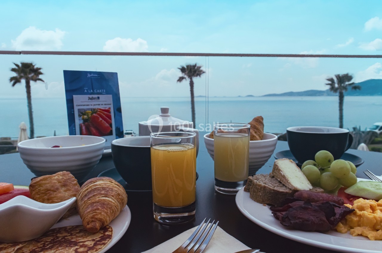 Petit-déjeuner varié avec fruits, viennoiseries, jus d'orange et vue sur la mer avec palmiers en arrière-plan.