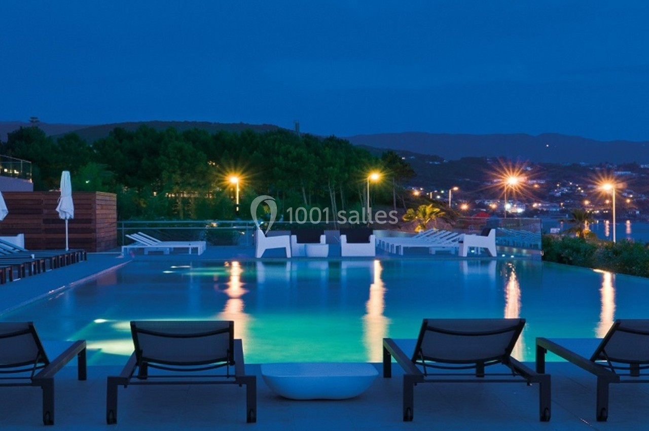 Piscine éclairée en soirée avec chaises longues au premier plan et vue sur une ville illuminée en arrière-plan.