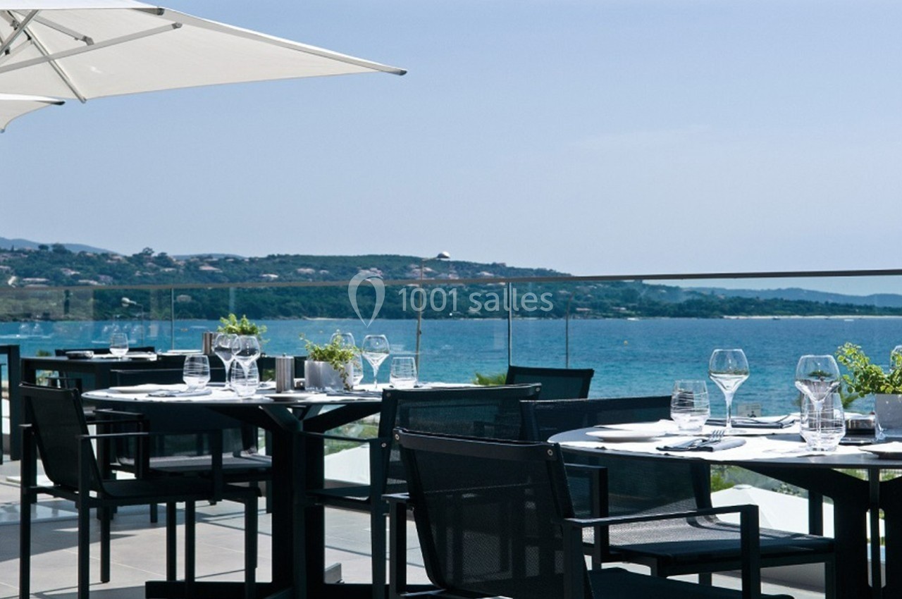Terrasse d'un restaurant avec tables dressées, vue sur la mer et collines en arrière-plan par temps ensoleillé.