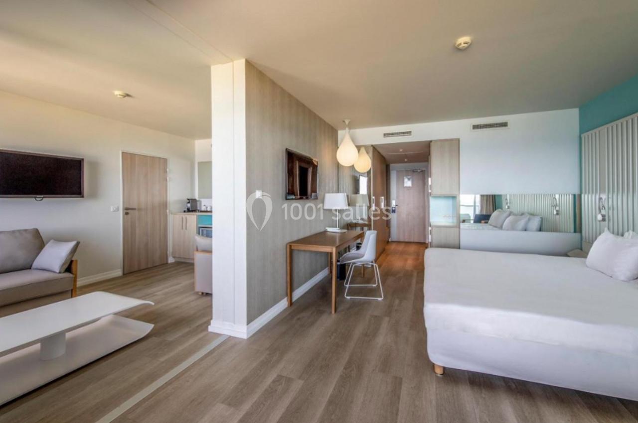 Chambre d'hôtel moderne avec lit double, coin salon, bureau, télévision murale et sol en parquet clair.