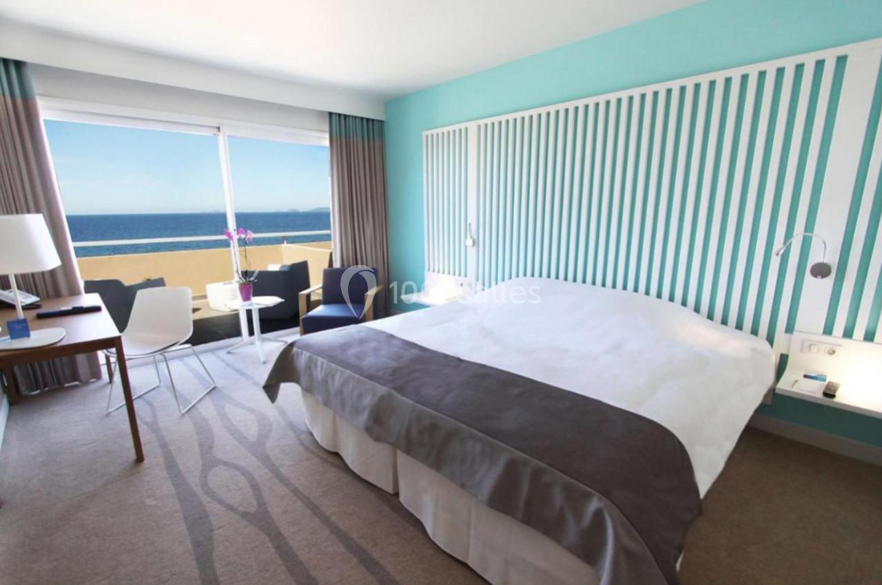 Chambre lumineuse avec lit double, bureau, fauteuils, baie vitrée donnant sur une terrasse et vue sur la mer.