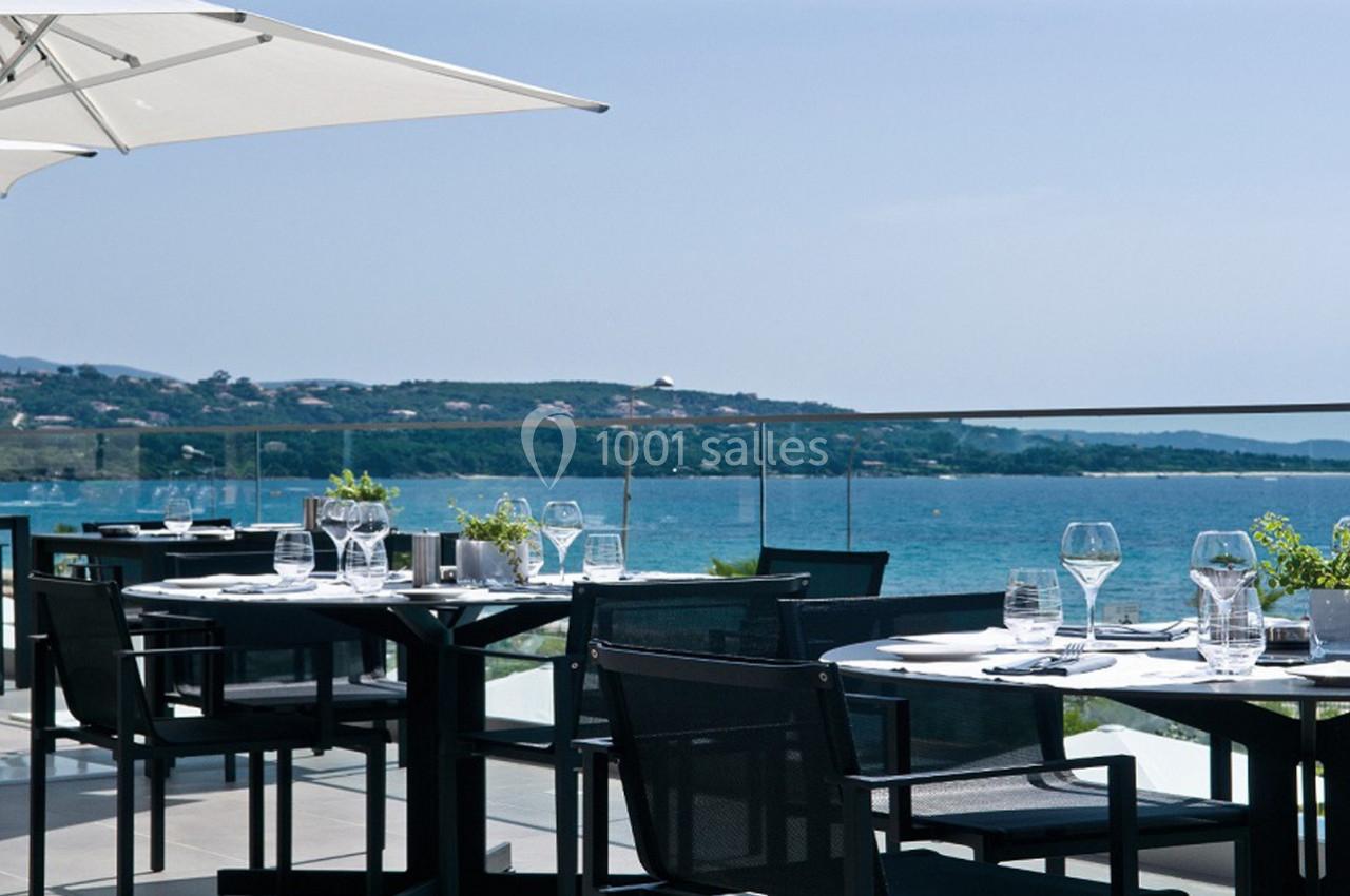 Terrasse d'un restaurant avec tables dressées, vue sur la mer et collines en arrière-plan par temps ensoleillé.