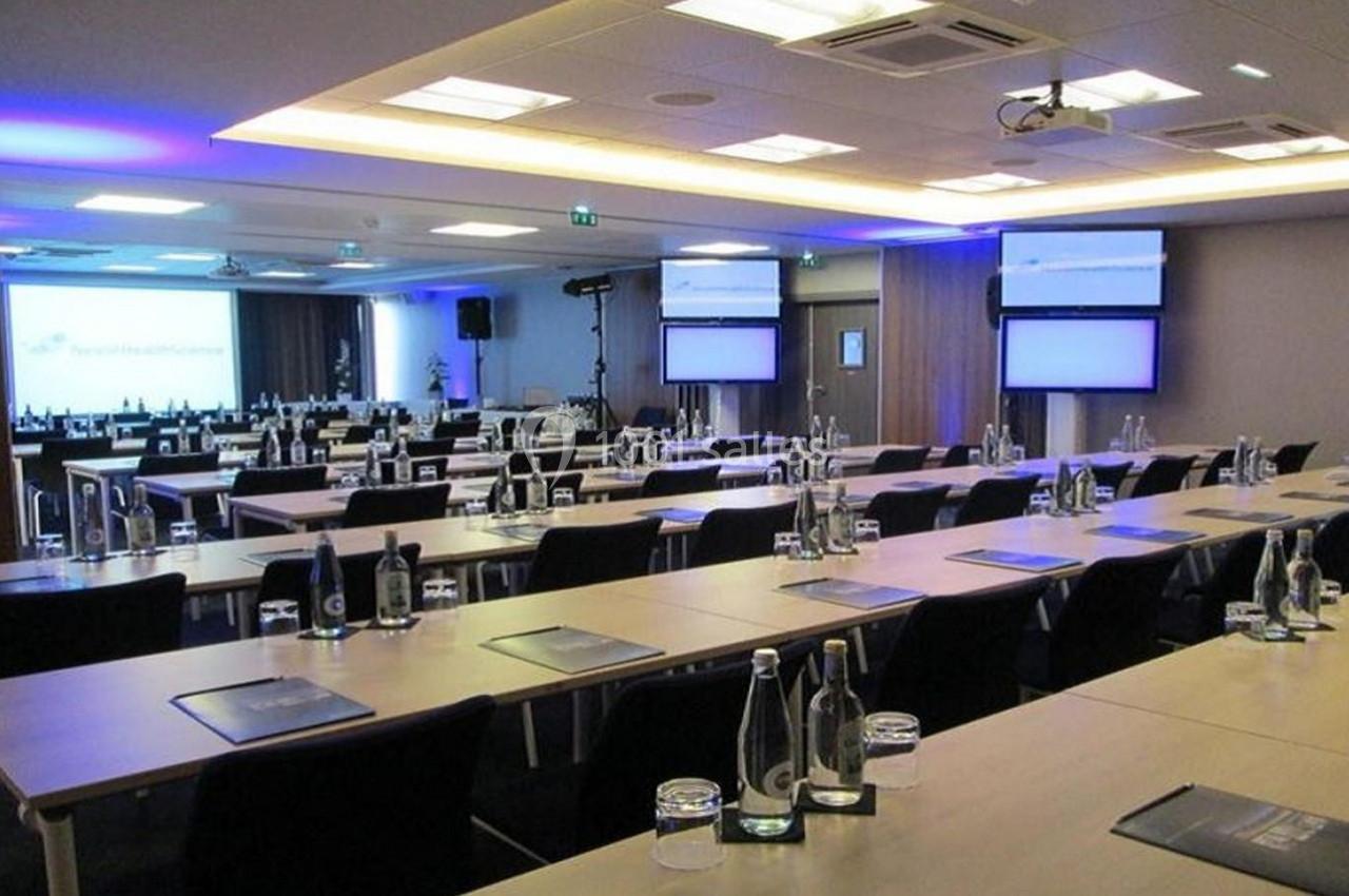 Salle de conférence moderne avec rangées de tables, chaises, écrans et bouteilles d'eau disposées pour un événement.