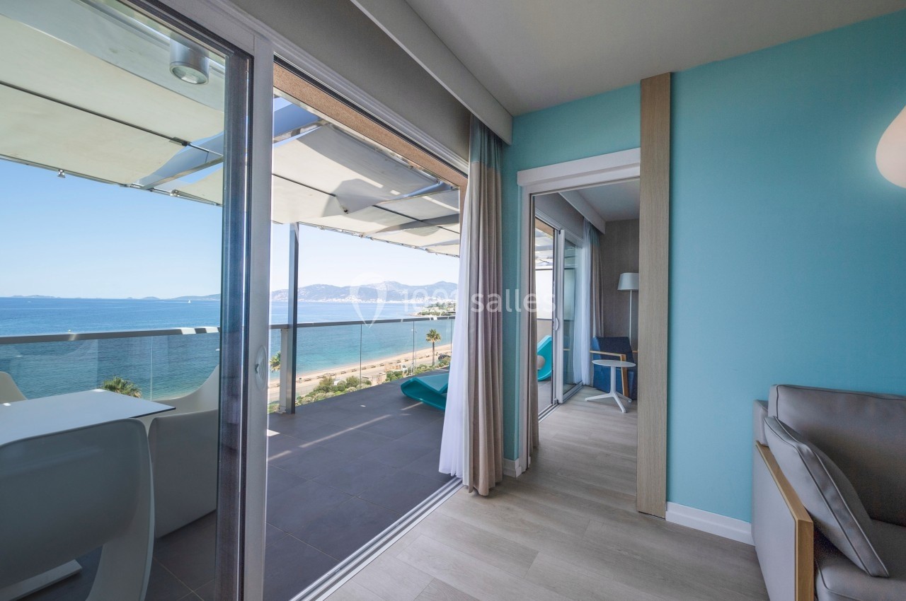 Vue depuis un salon lumineux sur une terrasse avec mobilier, donnant sur la mer et un paysage côtier ensoleillé.
