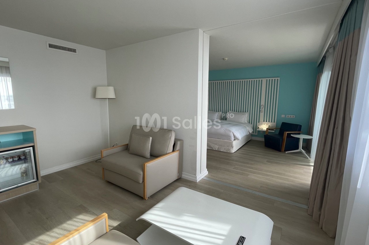 Chambre d'hôtel lumineuse avec coin salon, lit double, mini-réfrigérateur et grandes fenêtres laissant entrer la lumière…