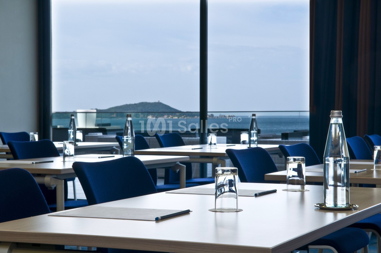 Salle de réunion lumineuse avec des tables alignées, des chaises bleues, des bouteilles d'eau et vue sur la mer.