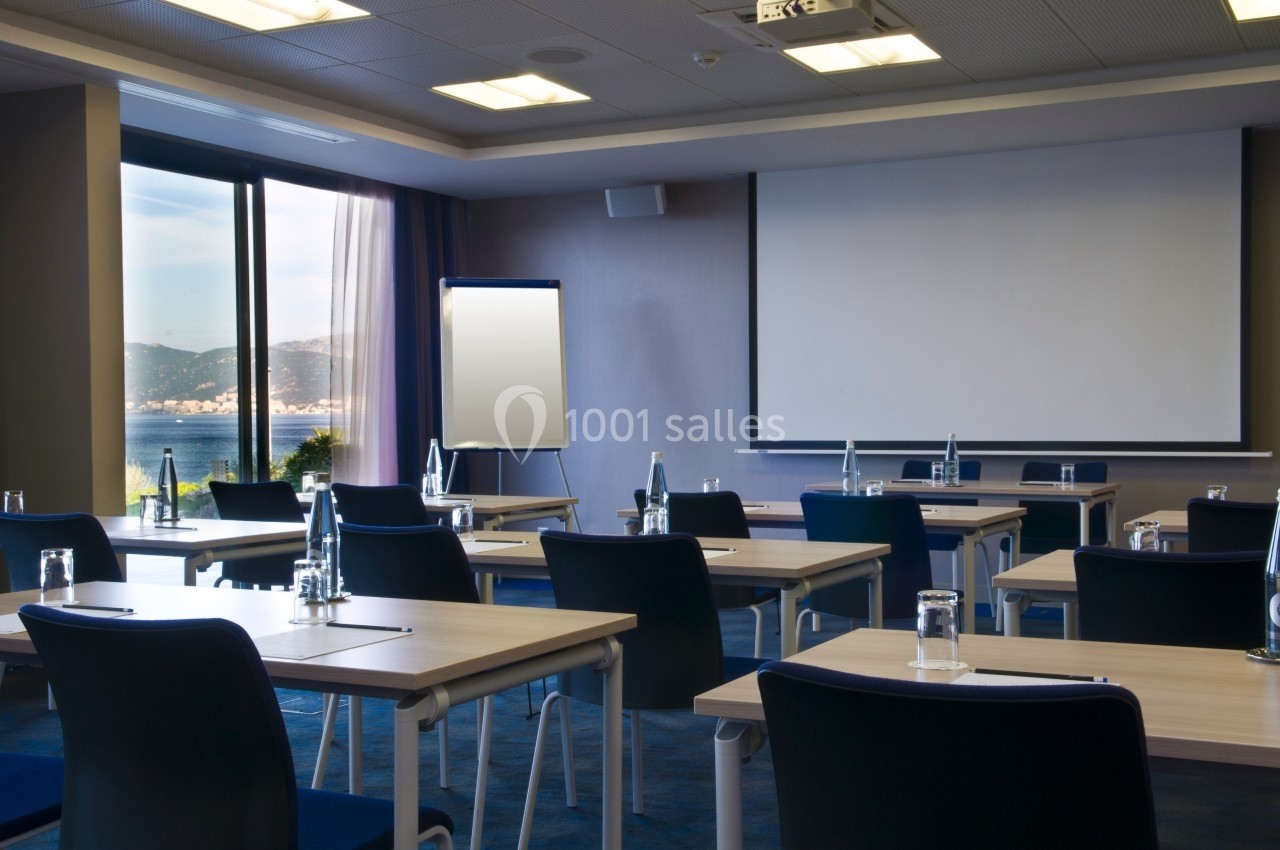 Salle de réunion lumineuse avec tables, chaises, écran de projection, tableau blanc et vue sur la mer.