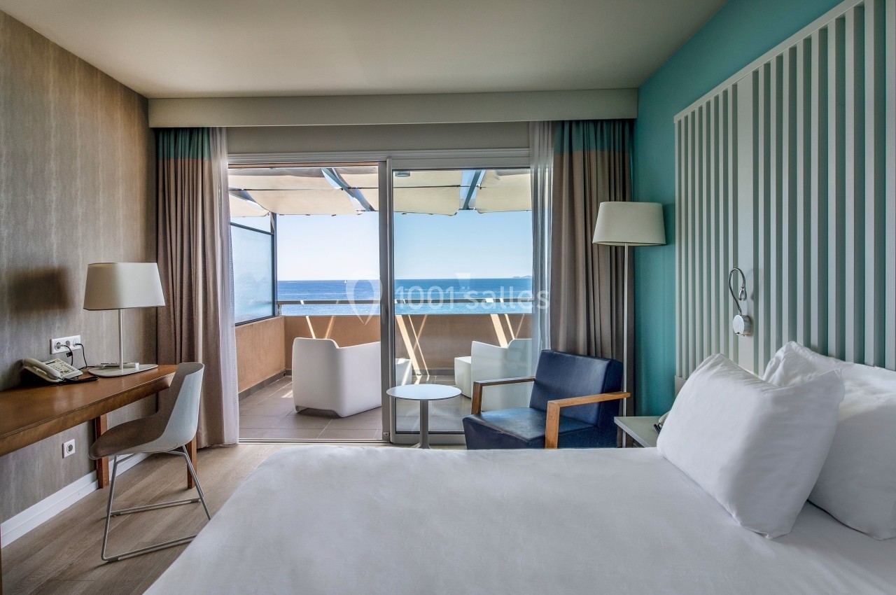 Chambre lumineuse avec lit double, bureau, fauteuils et balcon offrant une vue sur la mer.