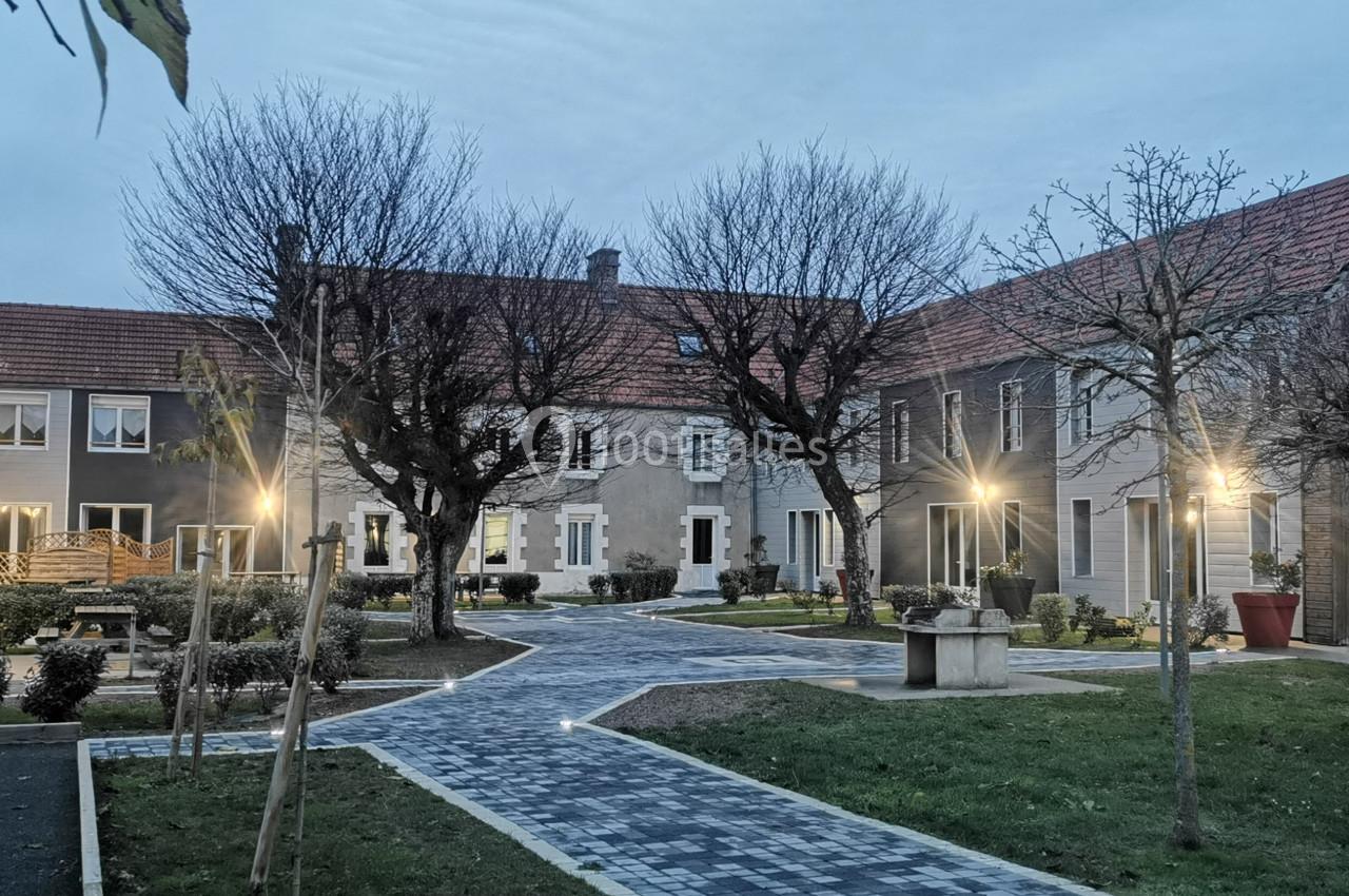 Bâtiment résidentiel entouré d'un jardin avec allées pavées, arbres sans feuilles et éclairage extérieur au crépuscule.
